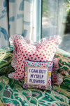 A decorative square pillow with the message "I CAN BUY MYSELF FLOWERS" in purple needlepoint text, set against a colorful floral border. The pillow is displayed on a green patterned sofa, in front of a larger ruffled pink pillow with a red and blue floral pattern, adding a cozy and eclectic touch to the decor.