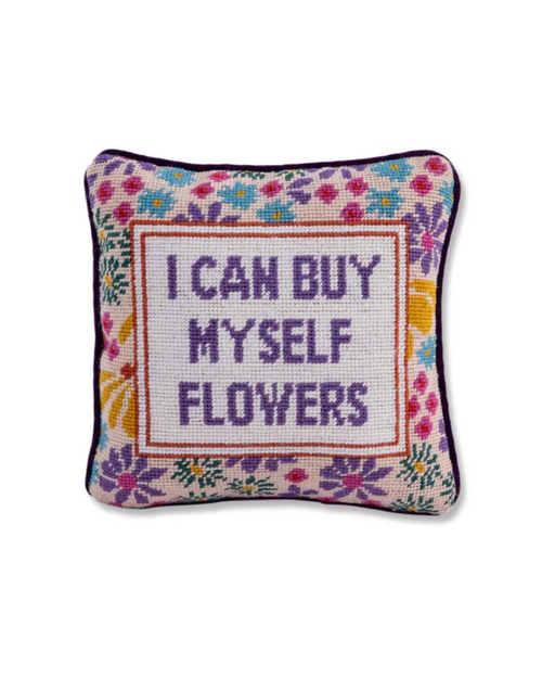 A square needlepoint pillow with a floral border in various colors. The center of the pillow has a stitched message in purple text that reads, "I CAN BUY MYSELF FLOWERS."