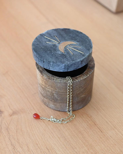 Image of a round moon box with the lid partially open, showing its interior and a chain inside. The lid features a brass moon and rays design. 