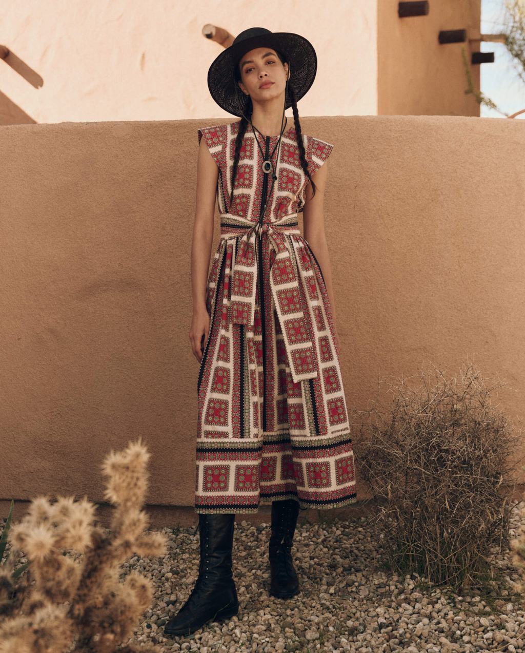 A woman in a desert setting wears a vintage-inspired print dress with a belt, paired with black lace-up boots and a wide-brimmed hat, conveying a rustic yet chic look.