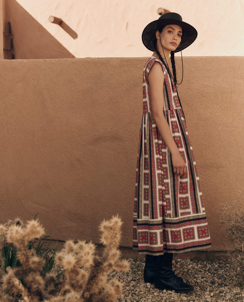 A woman in a desert setting wears a vintage-inspired print dress with a belt, paired with black lace-up boots and a wide-brimmed hat, conveying a rustic yet chic look.