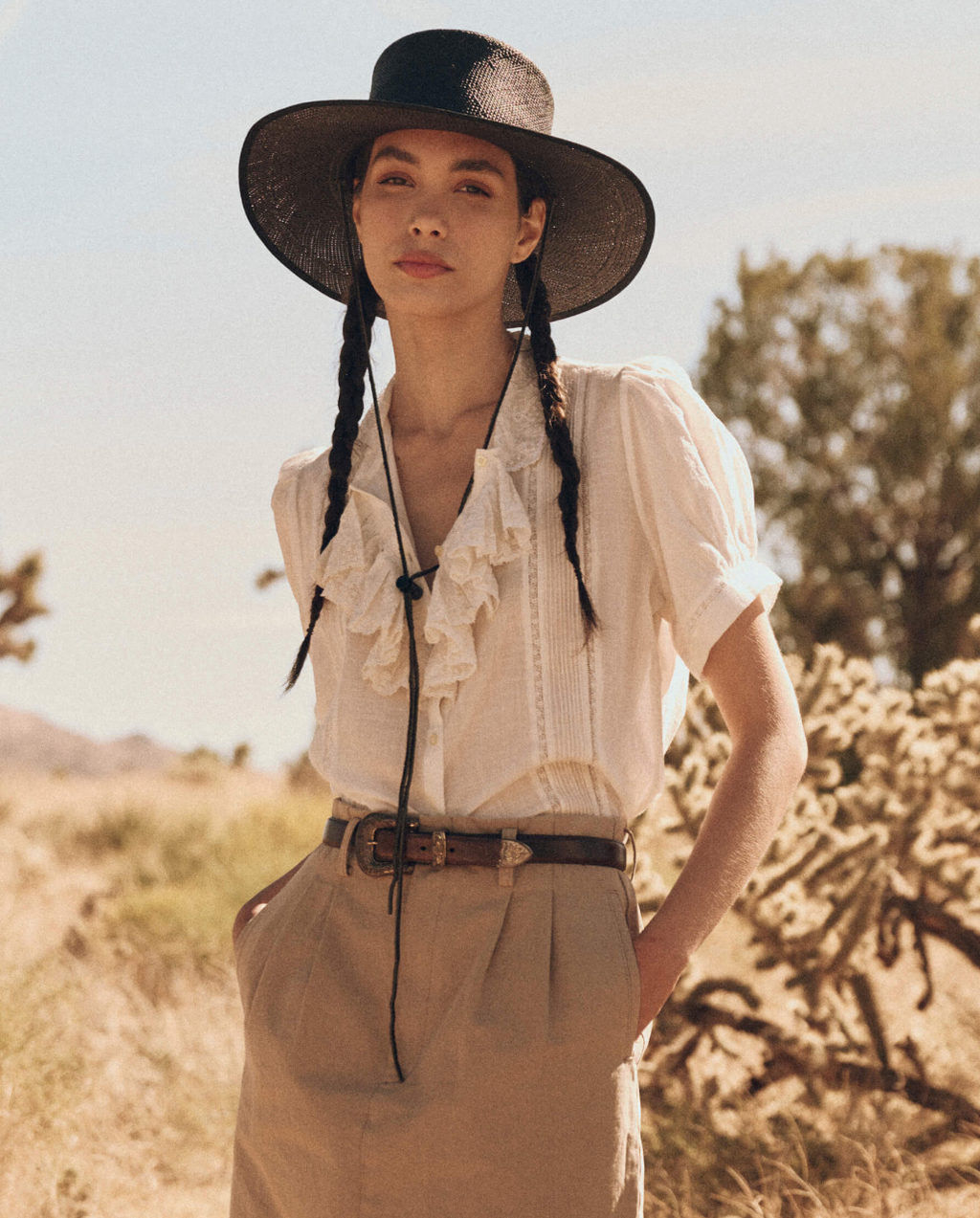 A woman wearing a Victorian-inspired top with delicate rose embroidery, dainty lace insets, and tiny pintucks. The top features an embroidered lace collar and a kerchief ruffle collar cascading down the front. She pairs it with high-waisted khaki pants and a wide-brimmed black hat with braids.