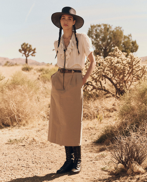 A full profile of a woman wearing a Victorian-inspired top with delicate rose embroidery, dainty lace insets, and tiny pintucks. The top features an embroidered lace collar and a kerchief ruffle collar cascading down the front. She pairs it with high-waisted khaki pants and a wide-brimmed black hat with braids.