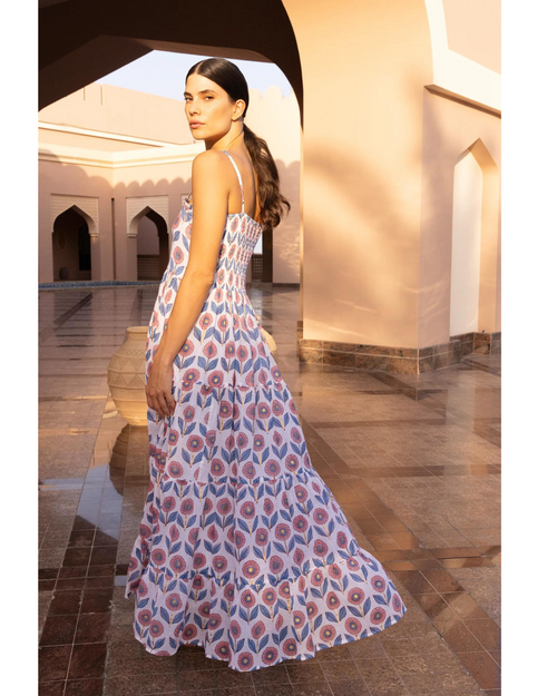 An elegant woman stands in a sunlit architectural setting, wearing a flowing maxi dress adorned with a vibrant pink and blue pattern. The dress features delicate straps and tiered ruffles, complementing her poised stance.