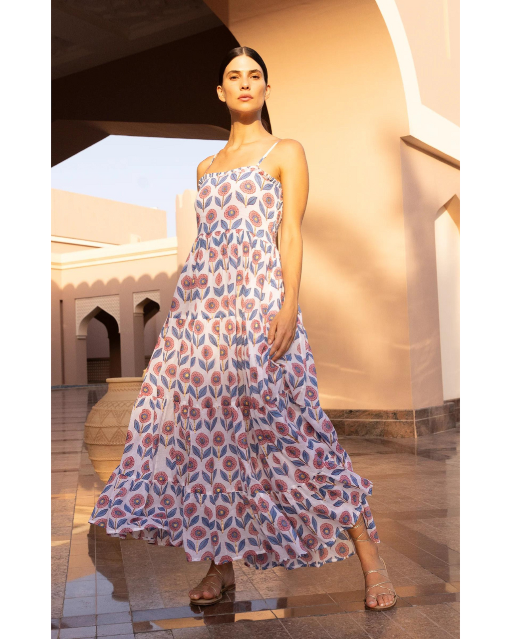 An elegant woman stands in a sunlit architectural setting, wearing a flowing maxi dress adorned with a vibrant pink and blue pattern. The dress features delicate straps and tiered ruffles, complementing her poised stance.