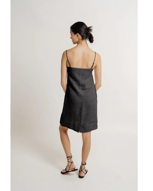 A model with a neat bun hairstyle is shown from the back, wearing the Square neck linen mini dress in black with adjustable straps, and black strappy sandals. The dress falls just above the knee.