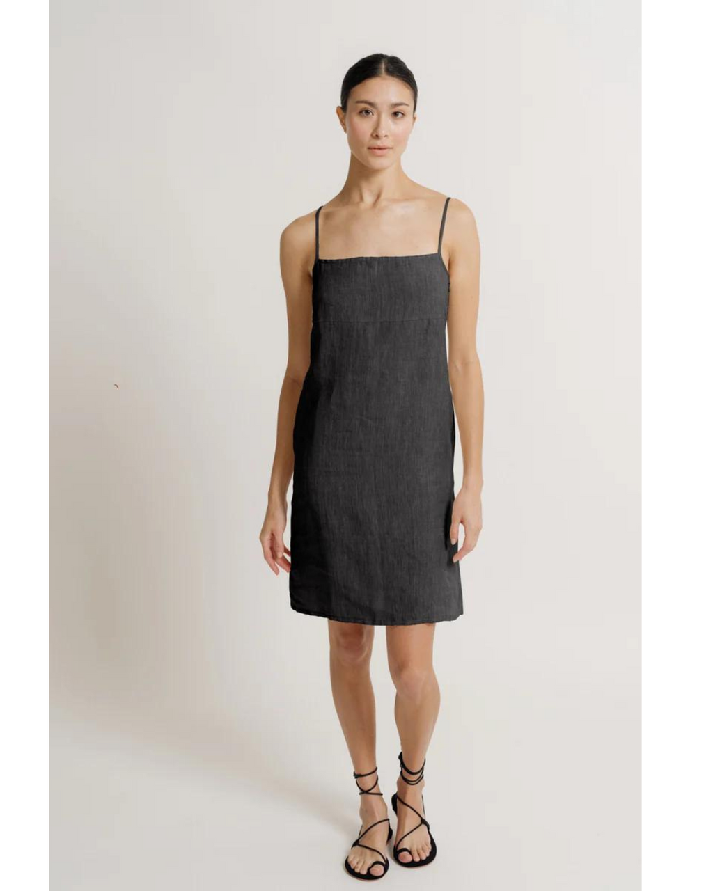 A model stands facing the camera, showcasing the front of a the Square Neck Linen Mini Dress in Black and adjustable straps. She pairs the dress with black strappy sandals.