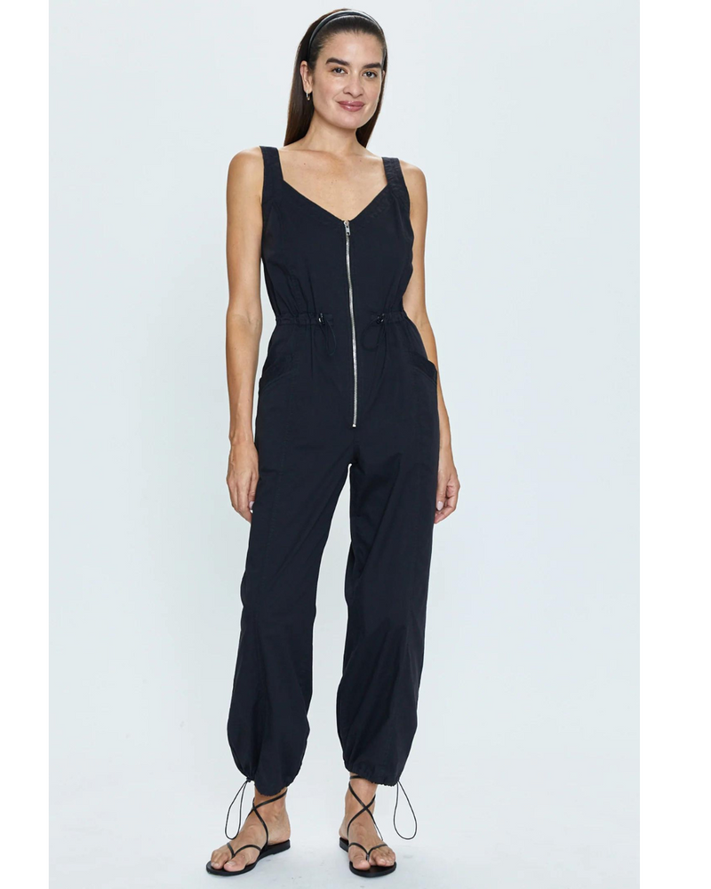 This image shows a woman modeling the Viva Parachute Tank Romper Noir. The romper features a sleek black color with a V-neckline and a visible front zipper. It has a tailored fit with adjustable waist and ankle ties for customization, complemented by side pockets for functionality. The model pairs the romper with thin-strapped black sandals, giving a chic and versatile look ideal for casual outings.