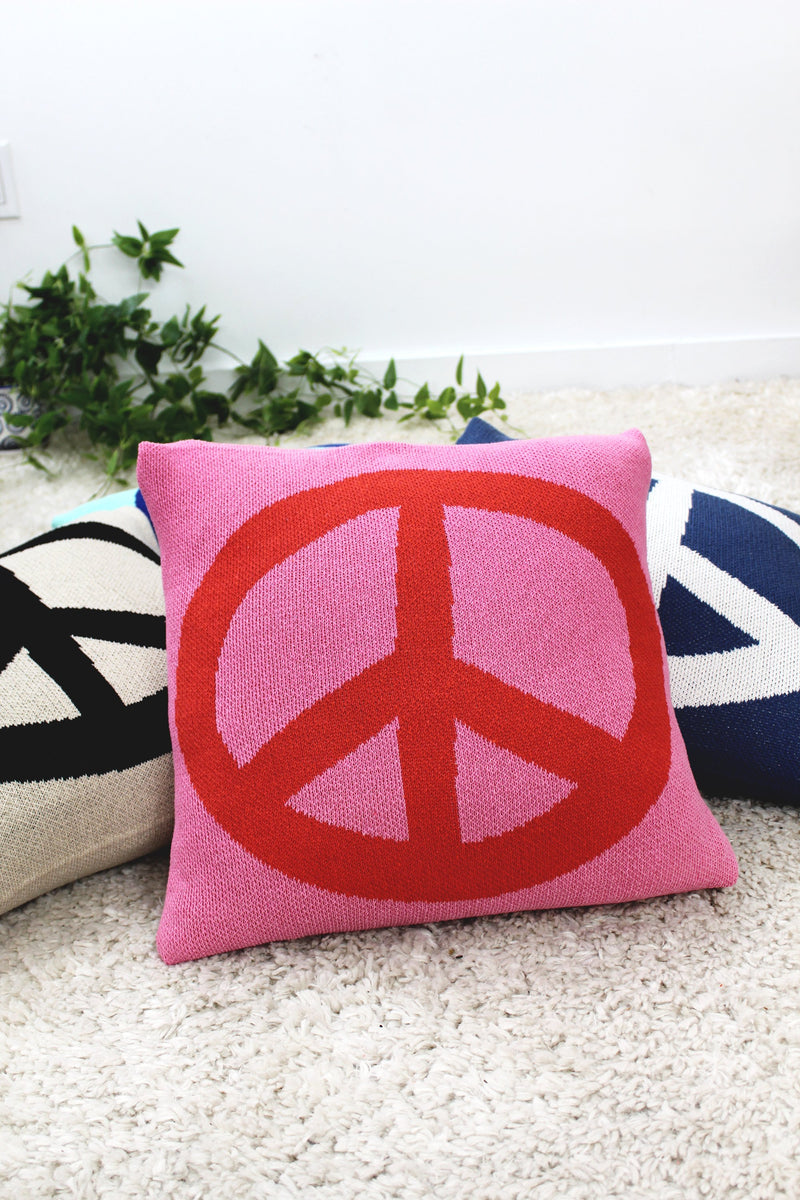 A pink pillow with a bold red peace sign design, surrounded by two other pillows featuring similar peace sign patterns in different color schemes. The pillows are displayed on a soft, beige carpet with green plant decorations in the background.