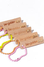 Five beige tags with the be golden logo engraved on them, each tag holding a different gold word stud earring: "Hey, Fuck, Yall, Love, Babe."