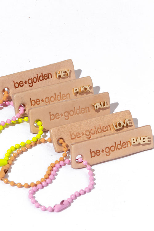 Five beige tags with the "be golden" logo engraved on them, each tag holdig a different gold word stud earring: "Hey, Fuck, Yall, Love, and Babe."