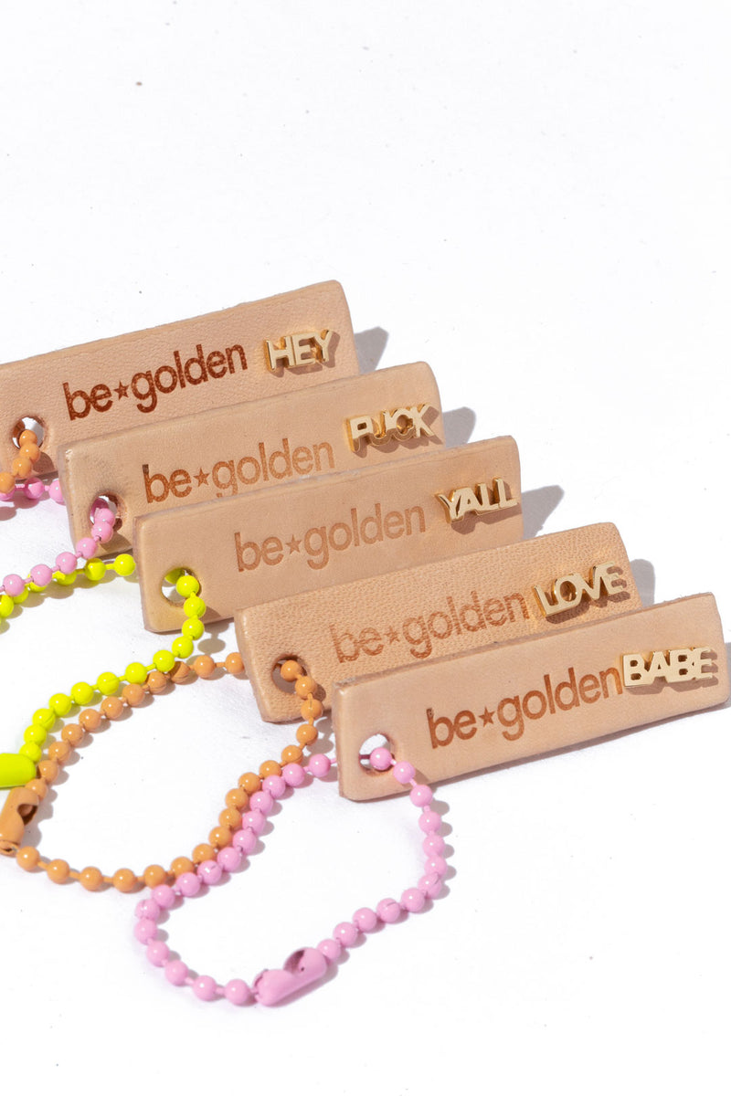 Five beige tags with the "be golden" logo engraved on them, each tag holdig a different gold word stud earring: "Hey, Fuck, Yall, Love, and Babe."