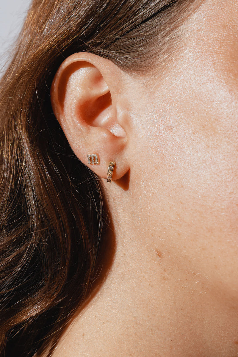 A close-up of a woman's ear wearing two gold earrings: a small, diamond-encrusted letter "m" stud in the upper earlobe and a small gold hoop earring with embedded diamonds in the lower earlobe. 