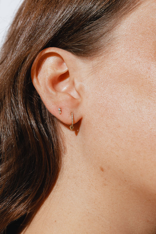 A close-up of a woman's ear wearing two gold earrings: a small, round stud in the upper earlobe and a gold hoop earring with a row diamonds and a pointed arrowhead top in the lower earlobe. 