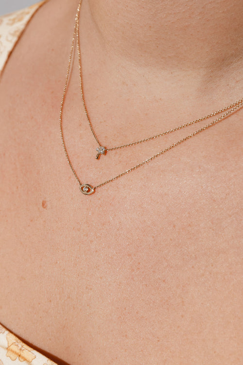 A close-up of a women's neck wearing two delicate gold necklaces. The shorter necklace features a small diamond-studded key pendant, while the longer necklace has an eye-shaped pendant with a central diamond. 