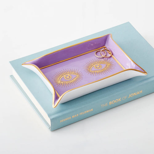 A rectangular tray with a purple interior and white exterior, featuring gold trim and two gold eye designs with radiating lines. The tray is placed on a light blue book titled "The Book of Jonah" by Joshua Max Feldman and holds two gold rings