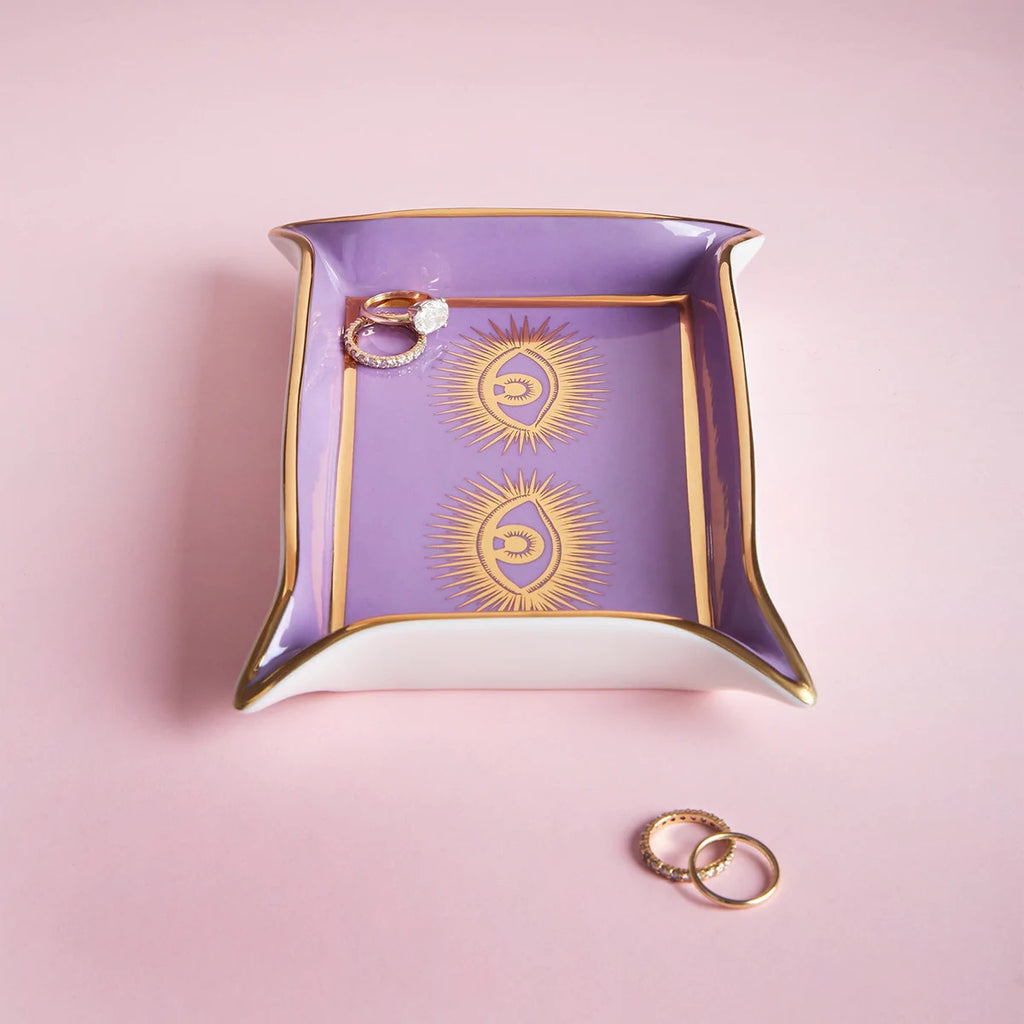 A rectangular tray with a purple interior and white exterior, featuring gold trim and two gold eye designs with radiating lines. The tray holds a gold ring with a large stone and is photographed on a pink background, with additional gold rings placed beside it.